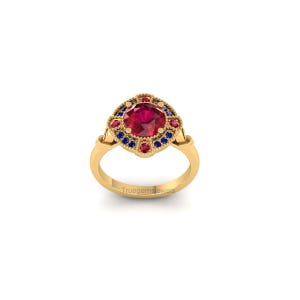 May include: A gold ring with a large, round, red gemstone at the center, surrounded by smaller red and blue gemstones. The ring has an ornate design and the text "truegemsjewels" is visible on the band.