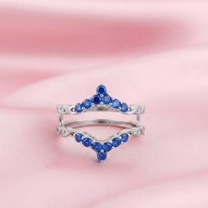 May include: A silver ring with a double band design. The ring features two rows of small, round, blue gemstones arranged in a chevron pattern. The ring is set against a soft pink background.