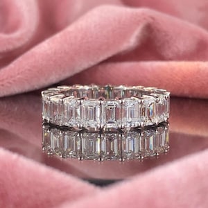 May include: A silver eternity ring with emerald-cut stones. The ring is set with multiple clear, rectangular-shaped stones. The ring is displayed on a reflective surface with a pink background.