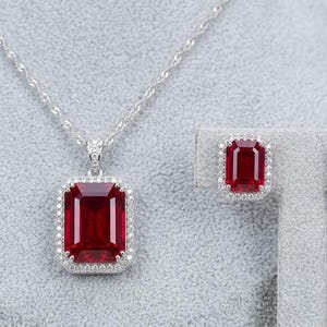 May include: A jewelry set featuring a pendant necklace and matching earrings. The pendant and earrings showcase large, rectangular, deep red gemstones, each surrounded by a halo of small, clear stones. The necklace has a delicate silver chain.