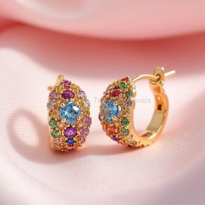 May include: A pair of gold-colored hoop earrings. Each earring features a central light blue gemstone surrounded by an array of colorful gemstones in various sizes and hues, including pink, purple, green, and orange. The earrings are set against a soft pink background.