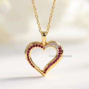 May include: A gold heart pendant necklace. The heart is edged with small red gemstones and white gemstones. The necklace chain is also gold. The pendant has the text "True Jewels" on it.