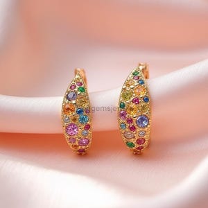 May include: A pair of gold-toned earrings, each adorned with a vibrant array of colorful gemstones. The earrings have a curved, leaf-like shape, with a stud post. The gemstones include shades of pink, blue, yellow, and green.