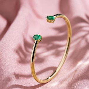 May include: A gold-colored open cuff bracelet with two oval-shaped emerald gemstones. The bracelet has a smooth, polished finish and a minimalist design. The gemstones are set in a bezel setting, adding a touch of elegance to the jewelry.