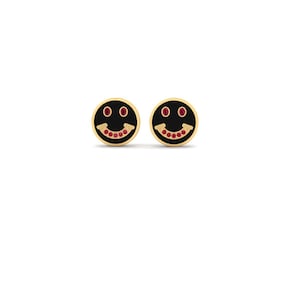 May include: A pair of gold-colored stud earrings. Each earring features a black circular face with a smiling mouth and two eyes, all outlined in gold. The mouth is filled with small red gemstones.