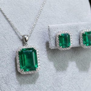 May include: A jewelry set featuring a pendant necklace and matching earrings. The pendant and earrings showcase rectangular, emerald-green gemstones surrounded by a halo of small, clear gemstones. The set is displayed on a gray surface.