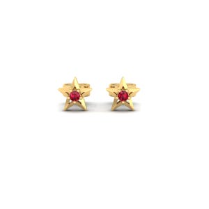 May include: A pair of gold-colored star-shaped stud earrings. Each earring features a five-pointed star design with a small, round, red gemstone in the center. The earrings are set against a white background.