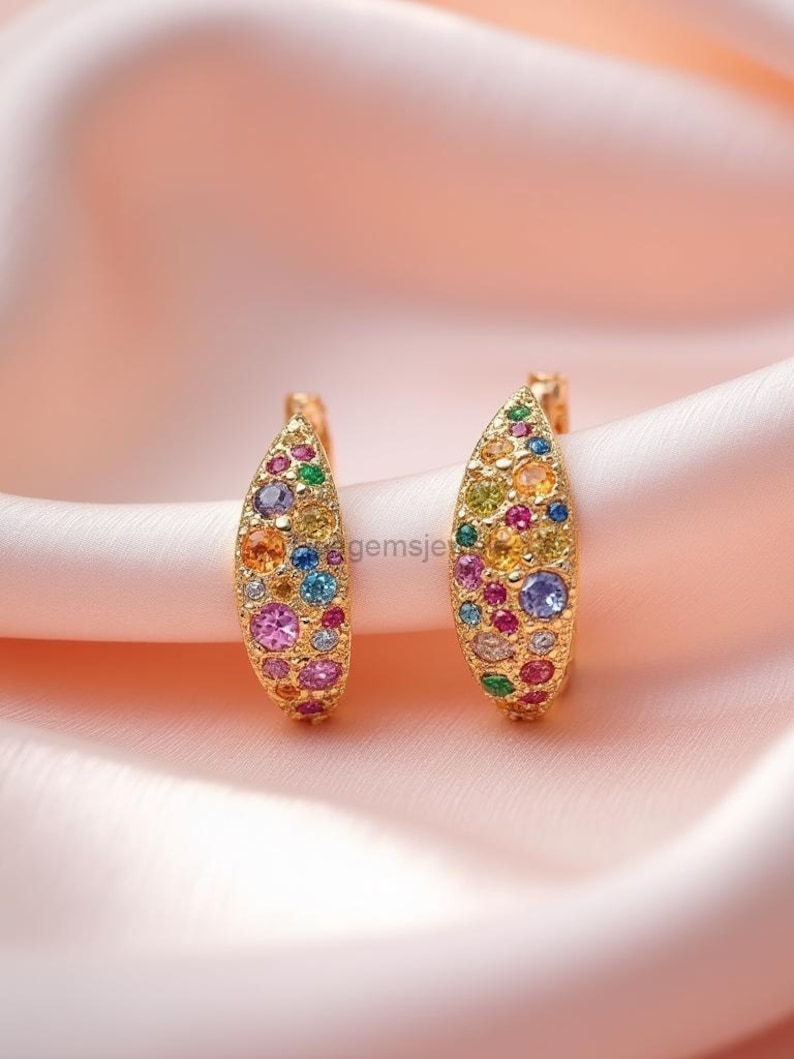 May include: A pair of gold-toned earrings, each adorned with a vibrant array of colorful gemstones. The earrings have a curved, leaf-like shape, with a stud post. The gemstones include shades of pink, blue, yellow, and green.
