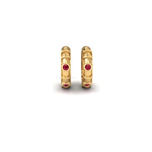 May include: A pair of gold hoop earrings, each with three red gemstones. The earrings have a textured design with vertical lines. The earrings are set against a white background.