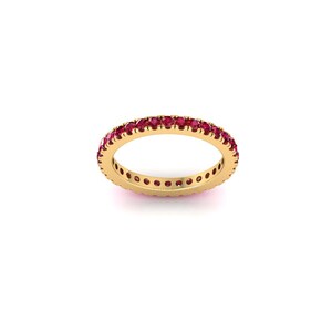 May include: A gold band ring set with numerous small, round, red gemstones. The ring is shown against a white background, highlighting the vibrant red of the stones and the warm tone of the gold.