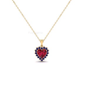 May include: A gold-colored necklace with a heart-shaped pendant. The pendant features a large, red heart-shaped stone surrounded by a halo of small, dark blue stones. The necklace is on a white background.