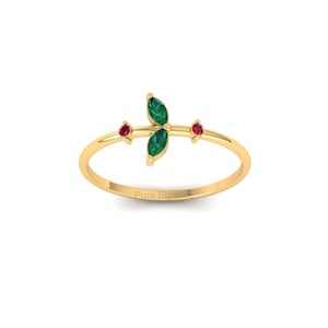 May include: A delicate gold ring featuring a leaf design with two emerald-colored stones and two small red stones. The ring has a thin band and is set against a white background. The text "Pure Elegance" is visible on the ring.