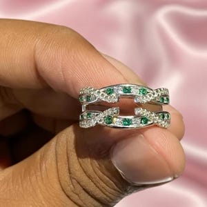 May include: A silver ring with a double band design. The bands are adorned with small, round, green and clear gemstones. The ring is held between fingers against a soft pink background.
