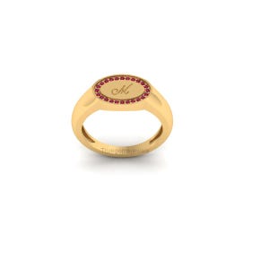 May include: A gold ring with an oval-shaped face. The face is bordered by small red gemstones and features the letter "M" in a script font. The ring is set against a white background.