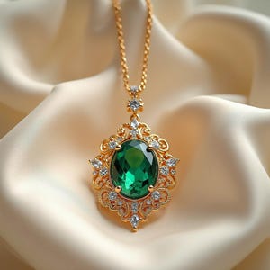 May include: A gold-toned necklace with a large, oval emerald-green gemstone pendant. The pendant is surrounded by a decorative gold filigree frame and small, clear crystals. The necklace chain is also gold.