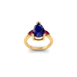 May include: A gold ring featuring a large, pear-shaped, deep blue gemstone as the centerpiece, flanked by two smaller, round, red gemstones. The ring has a classic, elegant design.