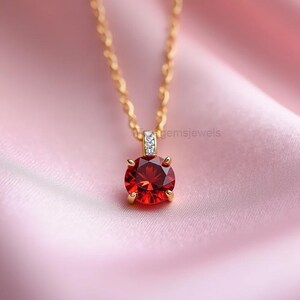 May include: A gold-colored necklace with a round, faceted, red gemstone pendant. The pendant is set in a gold-colored bezel and is accented with small, clear gemstones. The necklace is displayed on a pink satin background.