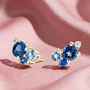 May include: A pair of gold stud earrings featuring clusters of blue gemstones in varying shades. The earrings have a sparkling, elegant design with a mix of dark and light blue stones and a small clear stone.