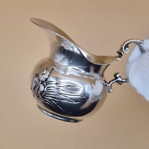 Puede incluir: Una pequeña jarra de crema plateada con un cuerpo redondeado y un pico curvo. La jarra de crema presenta un asa ornamentada y un diseño floral decorativo en la parte delantera. La superficie plateada refleja la luz.