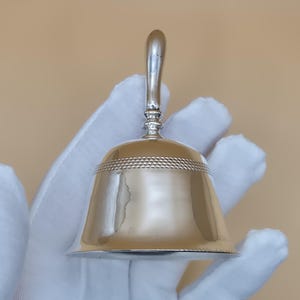 May include: A silver-colored bell with a rounded body and a curved handle. The bell has a decorative band of small, raised dots around its upper portion. The bell is held in a gloved hand against a neutral background.