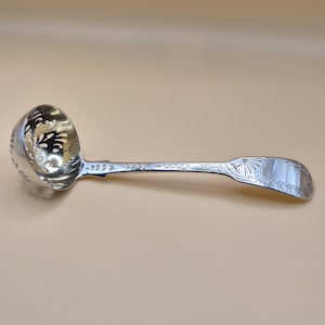 Könnte beinhalten: Eine silberfarbene Schöpfkelle mit einem runden, perforierten Löffel und einem langen Griff. Der Griff und die Schöpfkelle sind mit aufwendigen Blumen- und Blattmustern verziert. Die Schöpfkelle ist zum Servieren bestimmt und hat ein klassisches, elegantes Aussehen.