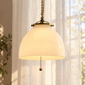 May include: A pendant light fixture featuring a smooth, white, dome-shaped glass shade attached to a brass-toned metal mount. A matching brass ball-chain pull switch hangs from the center, and the fixture is suspended by a thick, textured rope cord.