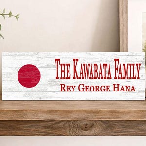 May include: A rectangular wooden sign with a white distressed finish featuring a red circle on the left side. The text in red reads The Kawabata Family followed by the names Rey, George, and Hana in a serif font centered on the right side.