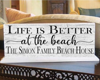以下が含まれることがあります： 「Life is Better at the beach」と「The Simon Family Beach House」という黒い文字が書かれた白い木製の看板です。