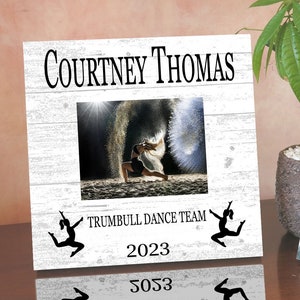 May include: White wooden picture frame with a white background. The frame has a photo of a dancer in the center. The text "Courtney Thomas" is at the top of the frame. The text "Trumbull Dance Team" is below the photo. The year "2023" is below the text. Two black silhouettes of dancers are on either side of the text.