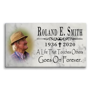 May include: A white marble memorial plaque with a black inscription that reads "ROLAND E. SMITH 1936† 2020 A Life That Touches Others Goes On Forever.". A black and white photo of a man wearing a hat is in the upper left corner.