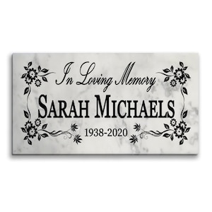 May include: A rectangular white marble plaque with black text reading In Loving Memory SARAH MICHAELS 1938-2020. The plaque features a black floral border design with leaves and flowers framing the text on the left and right sides.