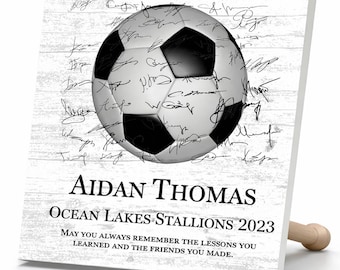 May include: A square white plaque with a wood grain texture featuring a black and white soccer ball graphic covered in signatures. The text reads: AIDAN THOMAS, OCEAN LAKES STALLIONS 2023, MAY YOU ALWAYS REMEMBER THE LESSONS YOU LEARNED AND THE FRIENDS YOU MADE.