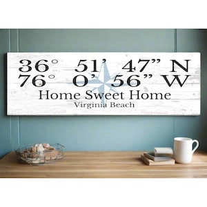 May include: White wooden sign with black text and a blue compass rose. The sign reads "36° 51' 47" N 76° 0' 56" W Home Sweet Home Virginia Beach".