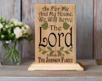 May include: A wooden sign with the text "As For Me And My House, We Will Serve The Lord" with a green leafy design and the text "Joshua 24:15" and "THE JOHNSON FAMILY".