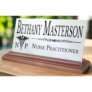 May include: White desk nameplate with black text and a medical symbol. The nameplate reads "Bethany Masterson, NP Nurse Practitioner". The nameplate is on a dark wood base.
