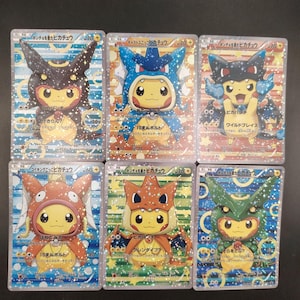 May include: Six holographic Japanese Pokemon trading cards featuring Pikachu wearing various costumes, including Umbreon, Gyarados, Charizard, Magikarp, and Rayquaza. Each card displays Japanese text, HP values, and attack statistics with a sparkling finish.