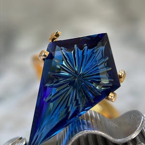 Puede incluir: Una piedra preciosa azul en forma de cometa con un patrón de facetas en forma de estrella, montada en una base de oro. La piedra muestra tonos azules profundos y claros, sujeta por cuatro garras de oro sobre una herramienta metálica ondulada.
