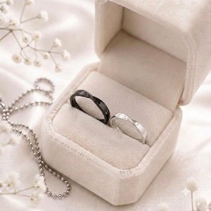 Op de afbeelding: Twee ringen, één zwart en één zilverkleurig, worden getoond in een beige fluwelen ringdoosje. De zilveren ring is gestempeld met S925. De ringen hebben een glad, gedraaid bandontwerp. Het doosje staat open en toont de ringen in de voering.