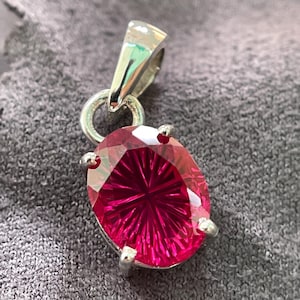 Op de afbeelding: Een hanger met een ovaalvormige, dieproze edelsteen met een radiale, zonnestraal-achtige geslepen facet. De steen wordt vastgehouden door vier zilverkleurige metalen pootjes en is bevestigd aan een gepolijst zilverkleurig metalen oogje.