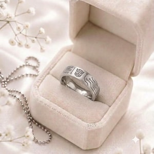 Puede incluir: Un anillo de plata con un símbolo Autobot grabado y un patrón de placa de circuito, marcado con S925. El anillo se muestra dentro de un joyero de terciopelo beige, acompañado de un collar de cadena de bolas plateada y flores blancas.