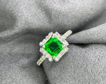 May include: A silver ring featuring a square-cut vibrant green center stone surrounded by a halo of small clear white stones. The band is thin and also set with small clear white stones along the visible portion of the metal shank.
