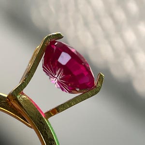 Peut inclure: Une bague en or avec une pierre pr&eacute;cieuse facett&eacute;e d'un rouge ros&eacute; profond, maintenue par un sertissage &agrave; griffes. La pierre pr&eacute;sente une taille brillant avec de multiples facettes r&eacute;fl&eacute;chissant la lumi&egrave;re, et l'anneau m&eacute;tallique est textur&eacute;.