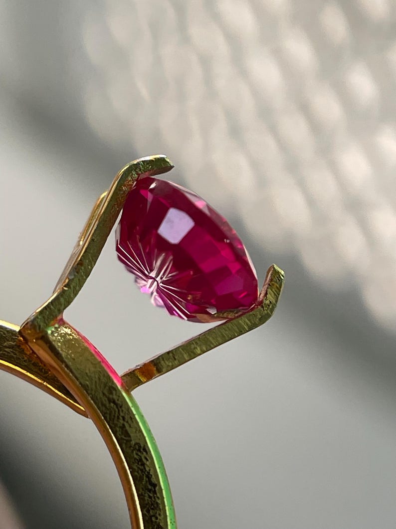 Peut inclure: Une bague en or avec une pierre pr&eacute;cieuse facett&eacute;e d'un rouge ros&eacute; profond, maintenue par un sertissage &agrave; griffes. La pierre pr&eacute;sente une taille brillant avec de multiples facettes r&eacute;fl&eacute;chissant la lumi&egrave;re, et l'anneau m&eacute;tallique est textur&eacute;.