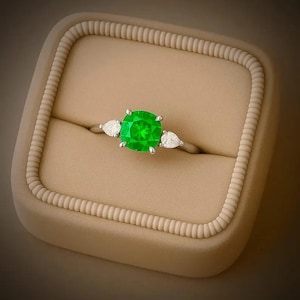 May include: A silver ring featuring a vibrant, square-cut, green gemstone at its centre, accompanied by two pear-shaped white stones. The ring is presented in a neutral-toned ring box.