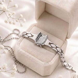 May include: A silver-toned metal bracelet featuring a central emblem of two overlapping wings, known as the Wings of Freedom, with a polished finish. The bracelet is stamped with S925 and rests inside a light-colored, velvet-lined jewelry box.