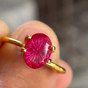 Peut inclure: Une bague en or orn&eacute;e d'une pierre pr&eacute;cieuse fuchsia de taille ovale avec un motif &agrave; facettes en &eacute;toile ou radial distinct. La pierre est fix&eacute;e par quatre petites griffes en or, et l'anneau est un fil de m&eacute;tal dor&eacute; lisse et poli.