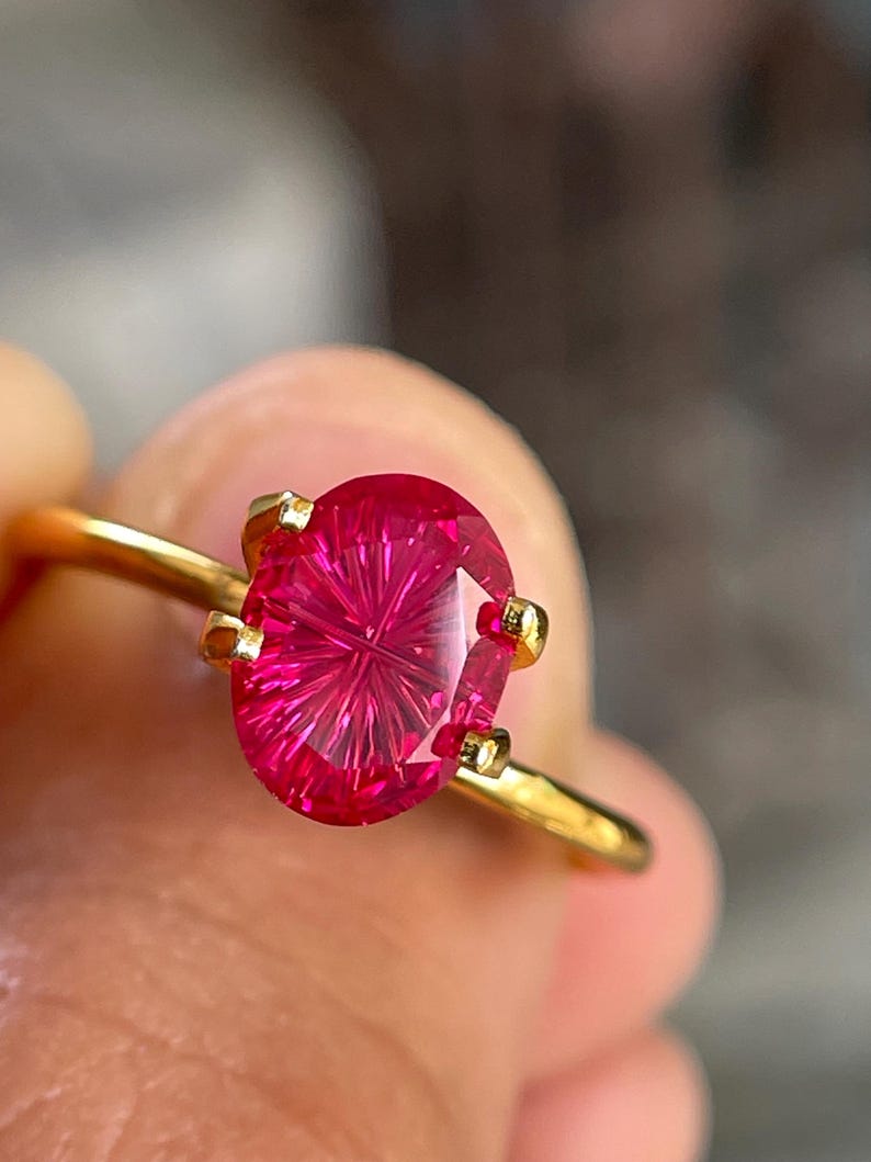 Peut inclure: Une bague en or orn&eacute;e d'une pierre pr&eacute;cieuse fuchsia de taille ovale avec un motif &agrave; facettes en &eacute;toile ou radial distinct. La pierre est fix&eacute;e par quatre petites griffes en or, et l'anneau est un fil de m&eacute;tal dor&eacute; lisse et poli.