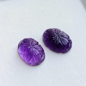 May include: Two oval-shaped, carved amethyst gemstones. The gemstones are a deep purple color with intricate floral designs carved into their surfaces. The carvings radiate from a central point, creating a starburst effect. The gemstones are displayed on a white surface.