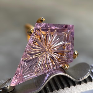 May include: A kite-shaped, light purple gemstone set in a gold ring. The stone features a complex, starburst-like faceted pattern radiating from the center. The ring is held by a metal tool with a serrated grip, showcasing the stone's clarity.