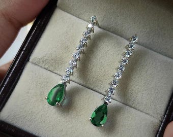 May include: A pair of elegant drop earrings. Each earring features a teardrop-shaped, vibrant green gemstone suspended below a linear arrangement of sparkling white gemstones. The earrings are set in a silver-toned metal and presented in a dark brown jewelry box.
